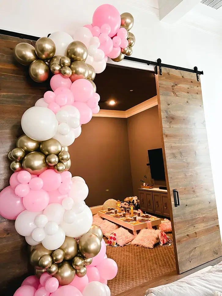 Guirnalda de globos rosa, blanco y dorado enmarcando la entrada a un salón con picnic