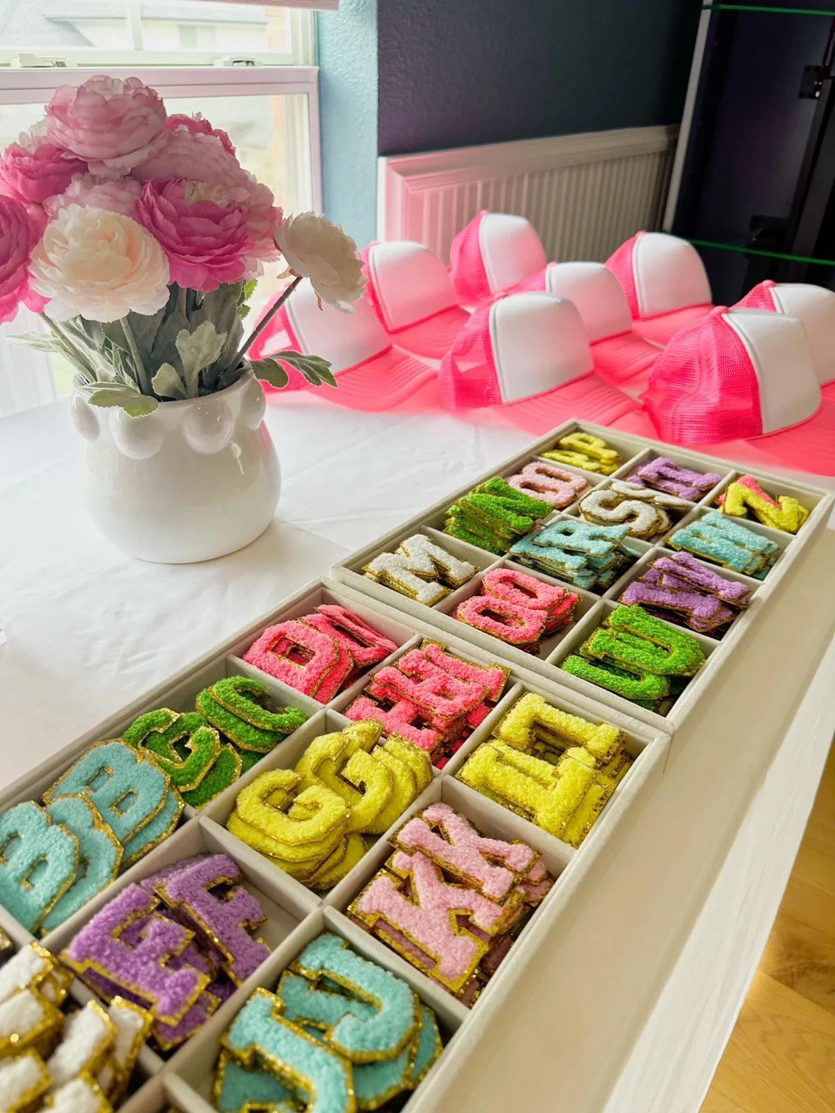 Pink trucker hats beside trays of colorful glitter letter patches
