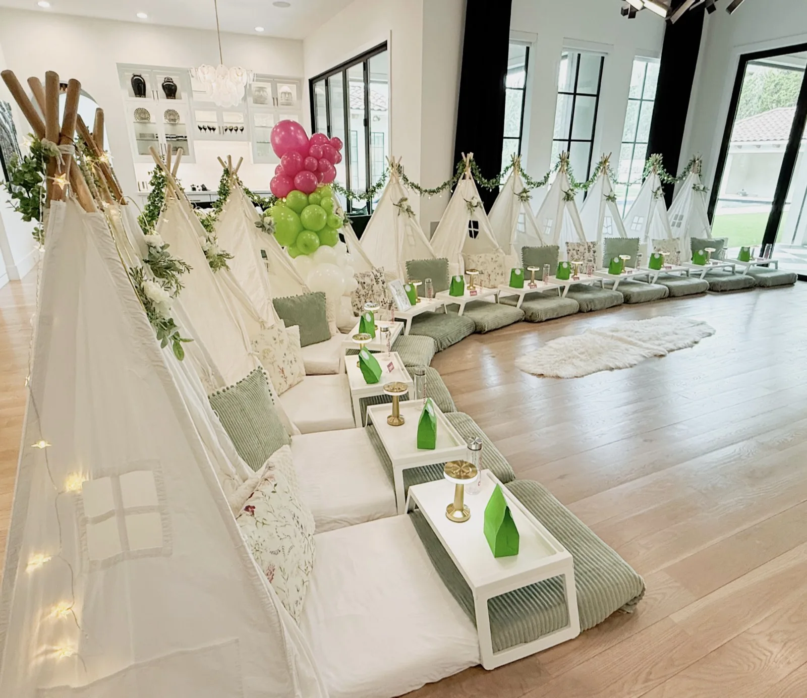 Una fila de teepees blancos con almohadones verde salvia, mesitas bandeja, faroles y una columna de globos rosa y verde en una sala luminosa